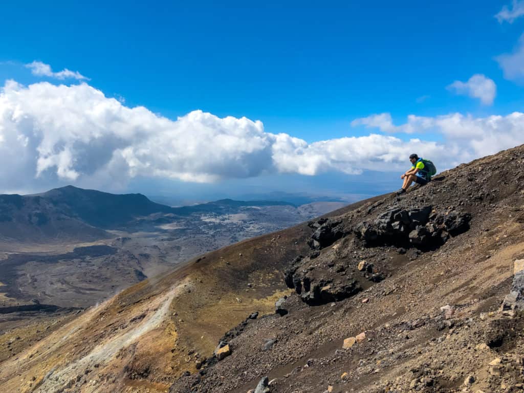 How to Hike the Tongariro Alpine Crossing and Why You'd Want To 8 Man sitting on the edge of the summit of the Tongariro Crossing with the clouds floating behind him