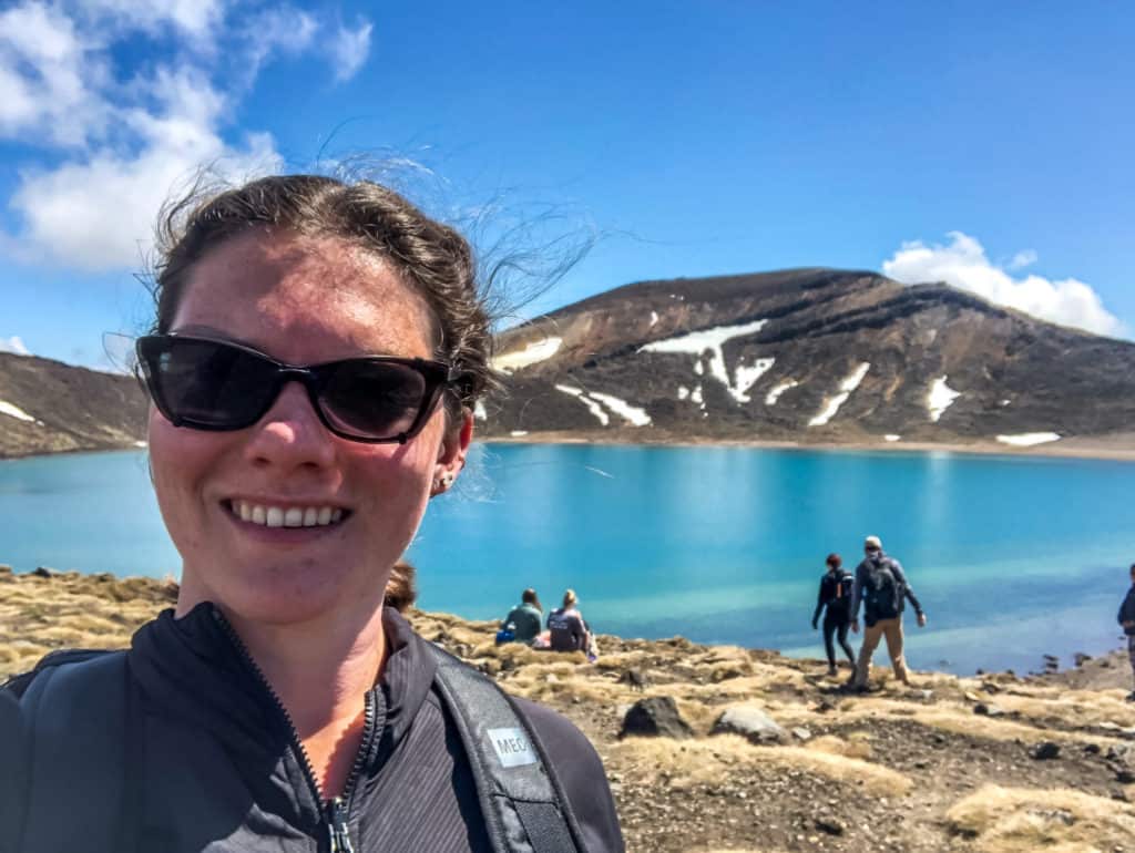 How to Hike the Tongariro Alpine Crossing and Why You'd Want To 12 Nina smiling in front of Mirror Lake at the Tongariro Alpine Crossing, with a snowy hill in the background.