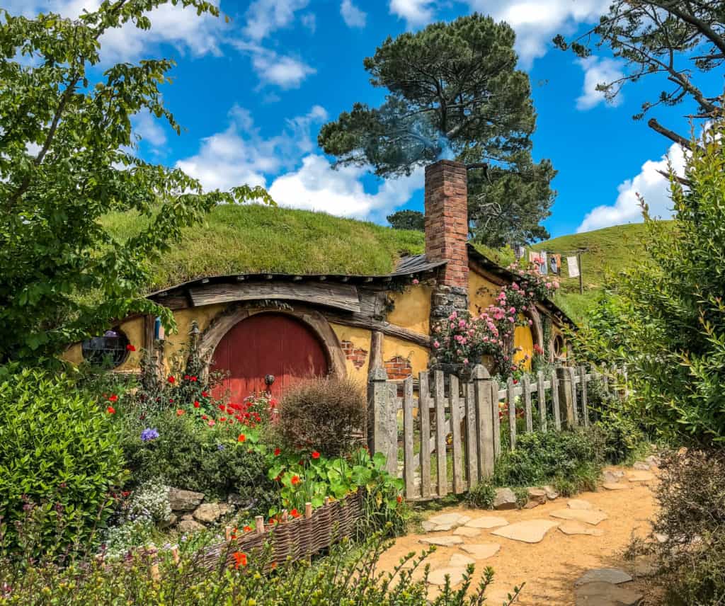 Visiting the Stunning Hobbiton Movie Set in New Zealand 2 Hobbiton hobbit hole with an ocher door and a chimney against the green of spring vegetation and a blue sky in New Zealand