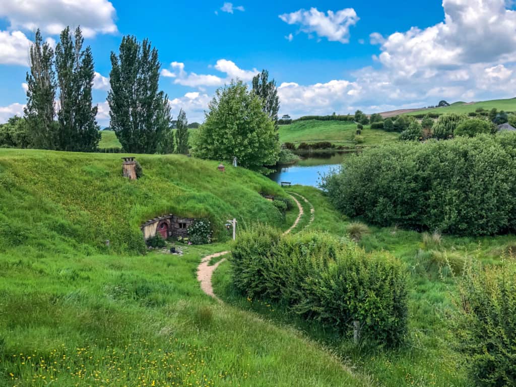 Visiting the Stunning Hobbiton Movie Set in New Zealand 3 A burgundy hobbit hole door set into the green grassy hillside of Matamata New Zealand with the blue lake and green trees surrounding it