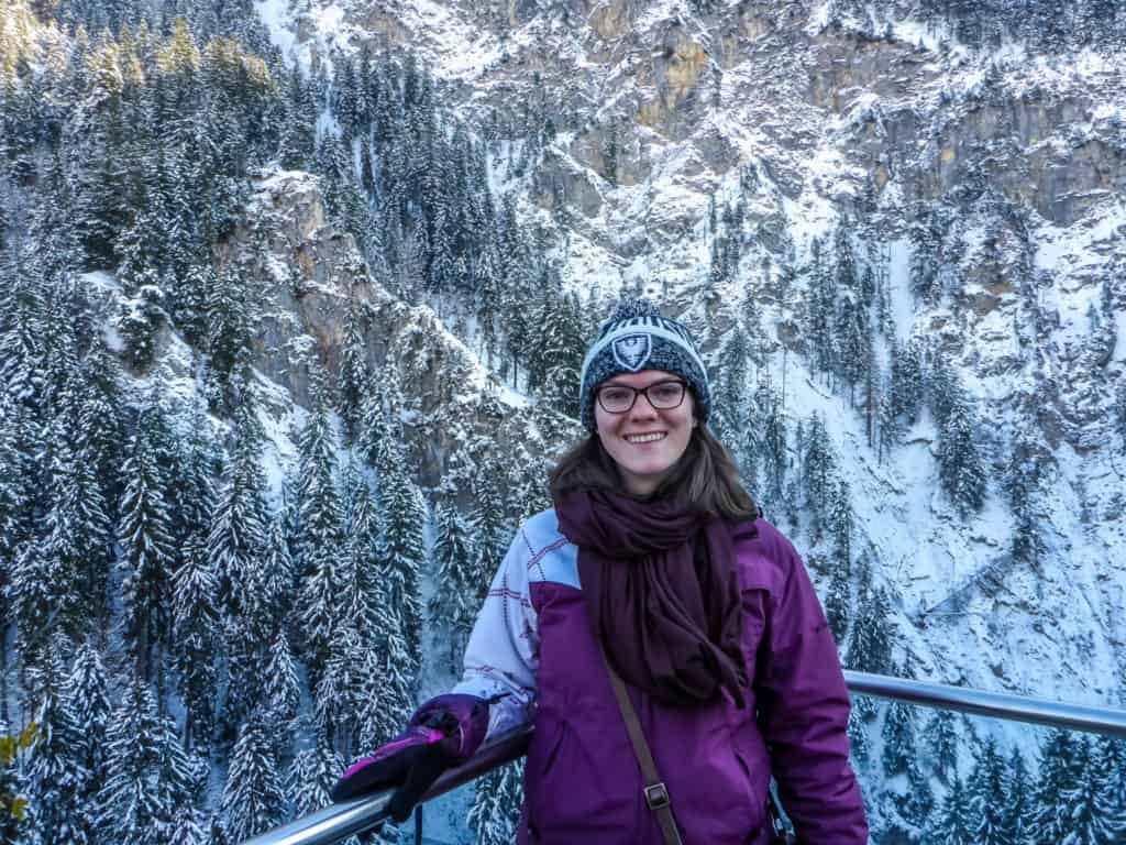 How to Visit Neuschwanstein Castle in Winter 2024 8 Nina smiling in a purple winter coat and grey beanie in front of the snowy Bavarian forest from the Neuschwanstein lookout in winter.