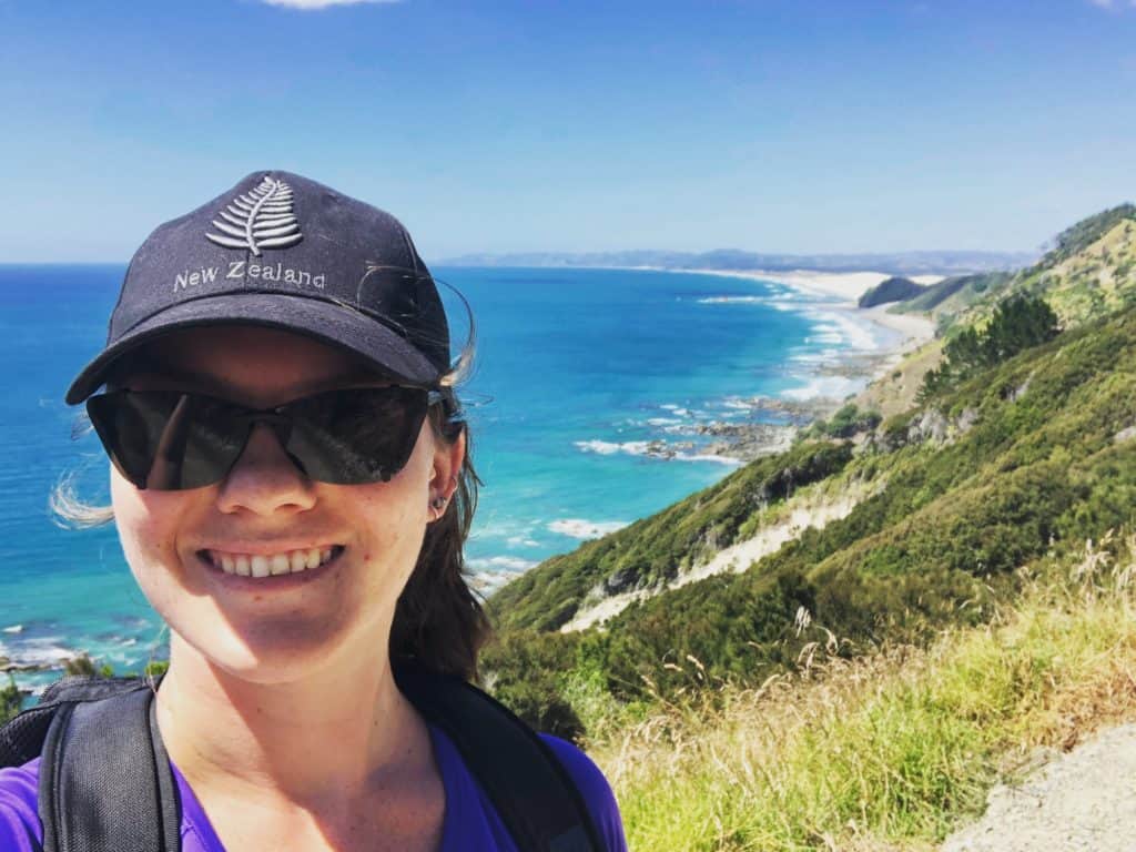 135 New Zealand Slang: From Sweet As to Swear Words 13 Nina wearing a New Zealand hat smiling with a background of the Mangawhai cliff walk of ocean and forests