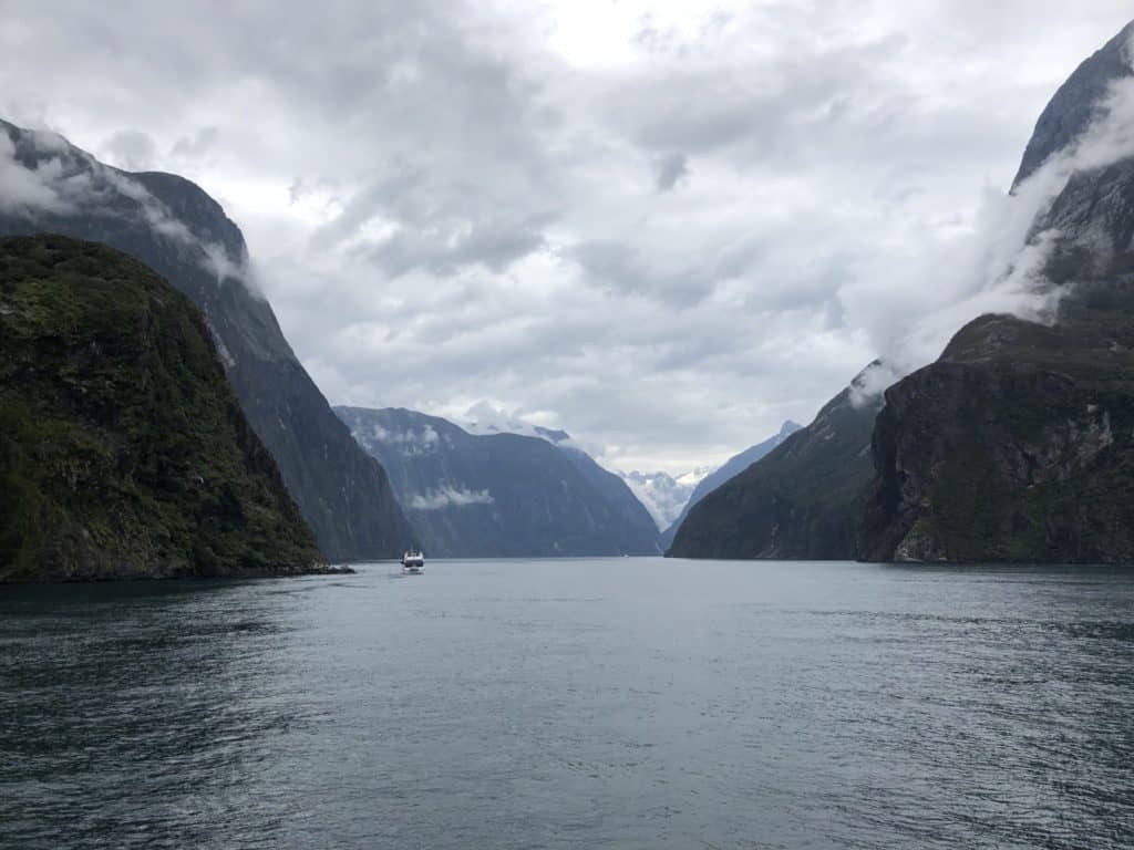 When is winter in New Zealand? (& Should You Visit?) 6 Milford sound with a boat in the distance on a cruise and the clouds hanging low over the sides of the fiord
