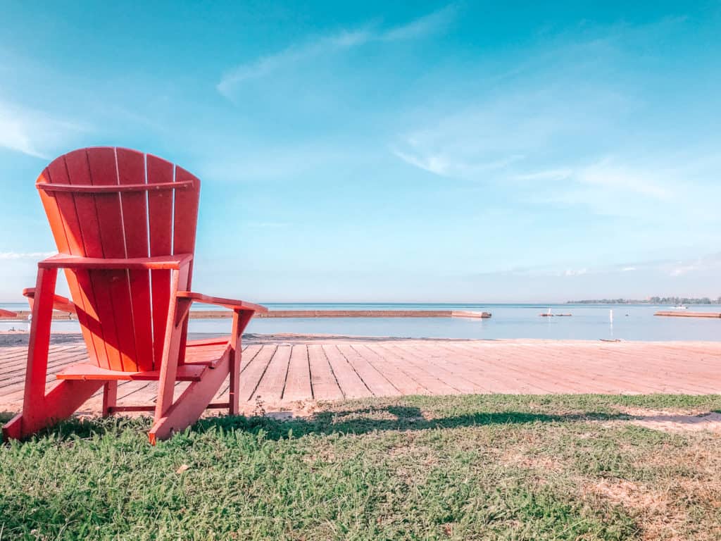 Is Toronto Worth Visiting in 2024? Local Guide 1 Red Muskoka chair on the Toronto boardwalk overlooking Lake Ontario and has a gorgeous clear blue sky above