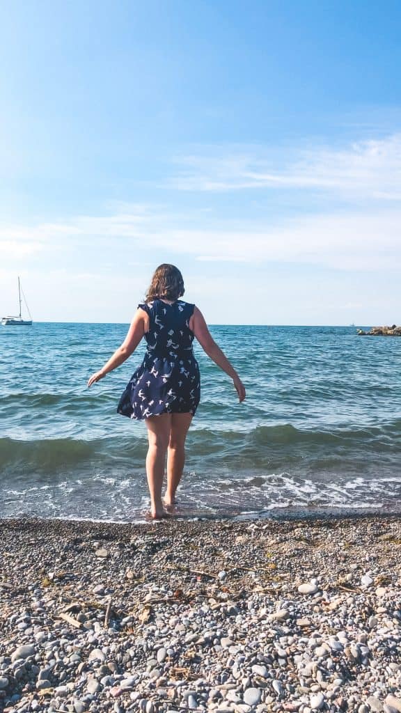 Is Toronto Worth Visiting in 2024? Local Guide 8 Nina wandering into Lake Ontario in the Beaches Neighbourhood in a blue dress with butterflies across it and a sailboat in the background in Toronto