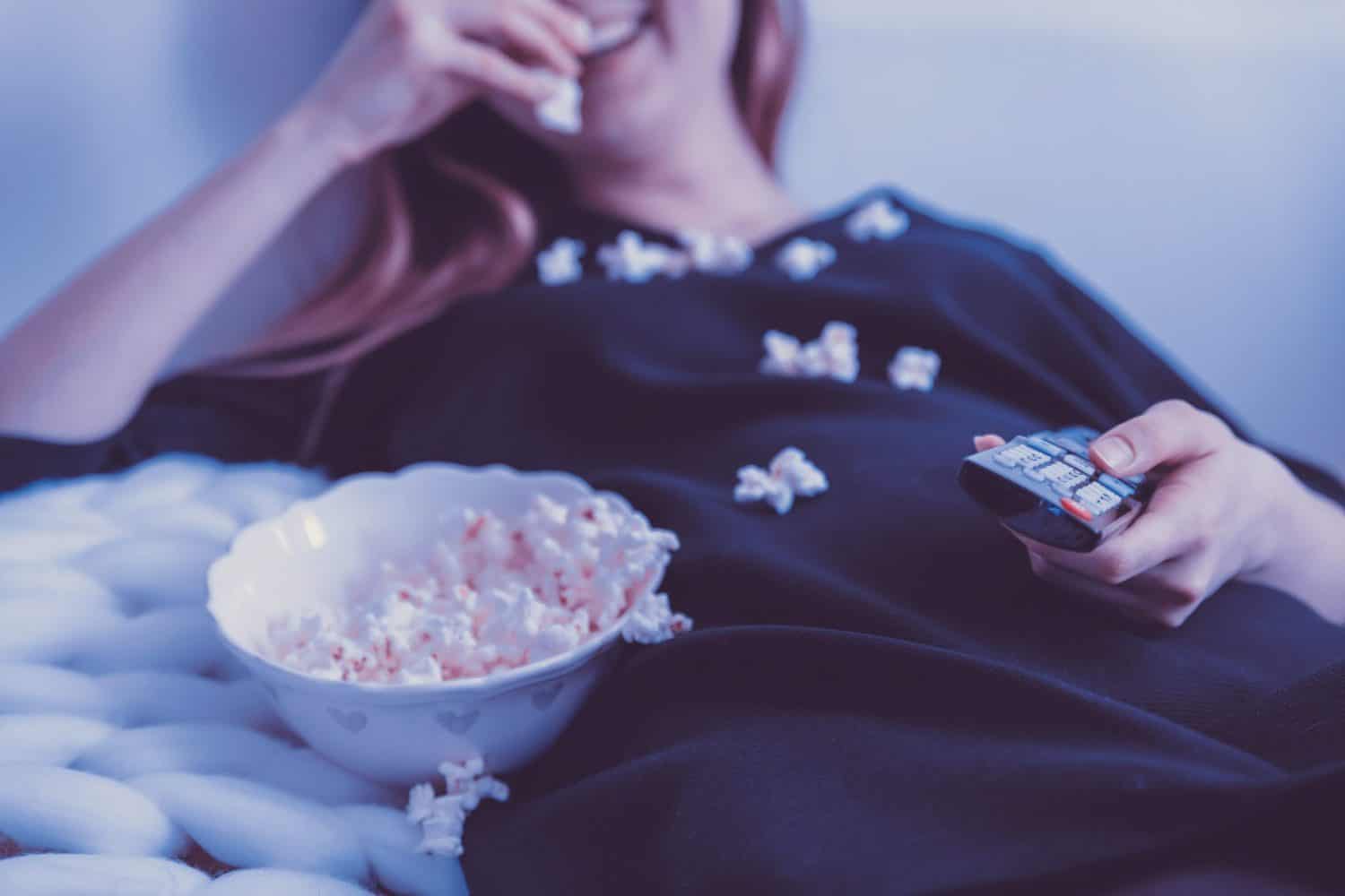 15 Unique Solo Staycation Ideas for Singles 12 Woman laughing eating popcorn while laying dying and holding a remote
