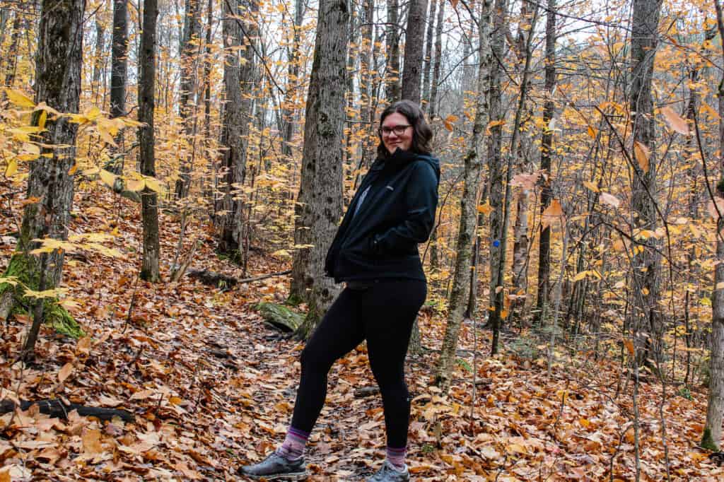 What to Wear Hiking: A Complete Guide for Beginners 5 Nina posing in hiking clothing layers in a forest of yellow leaves. Wearing a fleece lined rain jacket, leggings, hiking socks and sneakers.