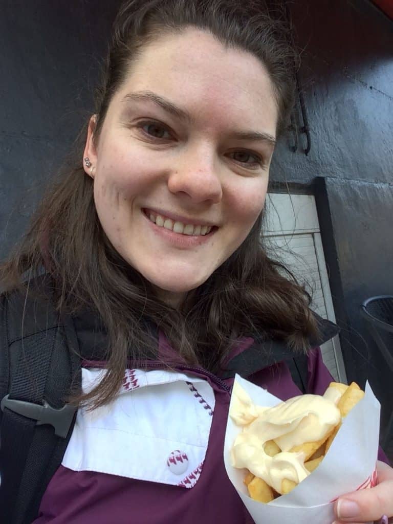 What You Need to Know Before Travelling to Amsterdam Alone 5 Nina smiling at the camera in a purple coat while holding a paper cone of french fries when she travelled to Amsterdam alone