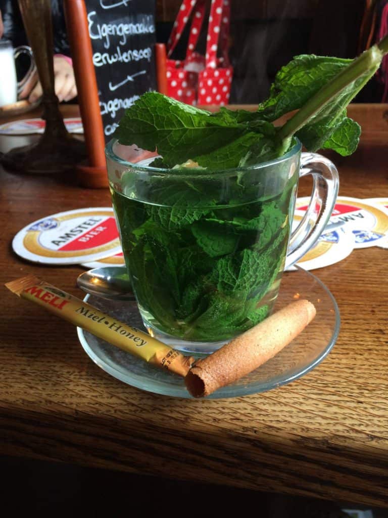 What You Need to Know Before Travelling to Amsterdam Alone 4 Fresh mint leaves in a glass mug with a packet of honey and a rolled biscuit on the edge of the small plate beneath it