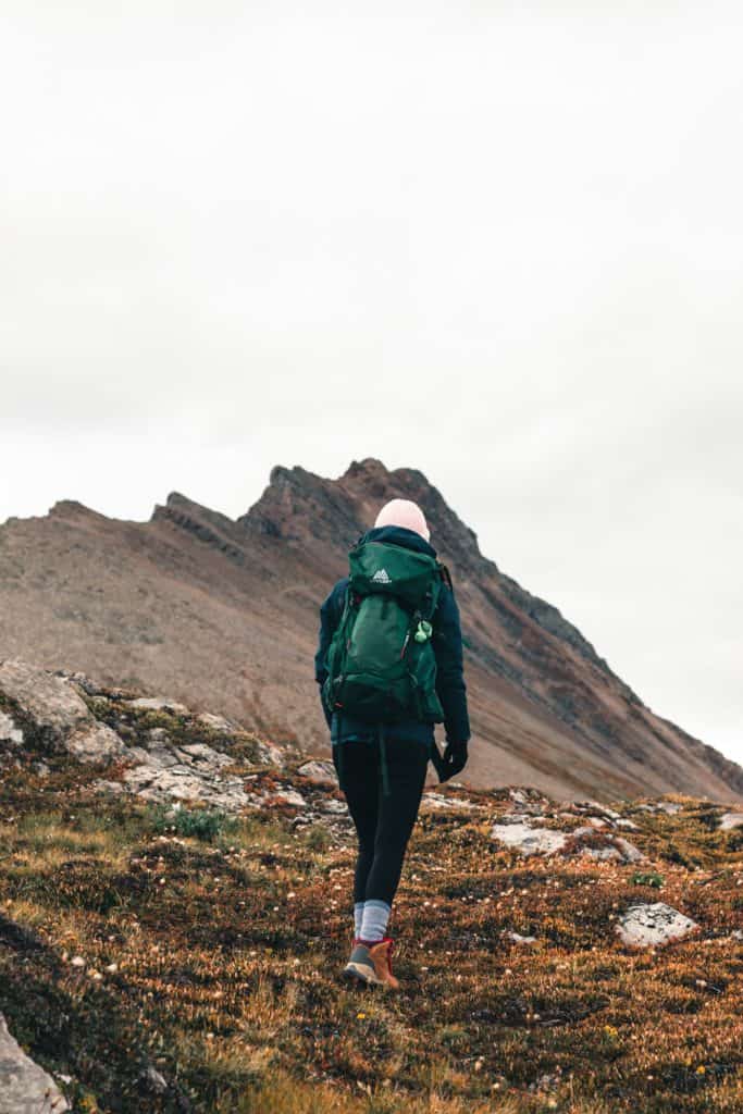 What to Wear Hiking: A Complete Guide for Beginners 3 Woman hiking a mountain climbing with lots of layered clothing, mittens, a hat, and a green backpack.