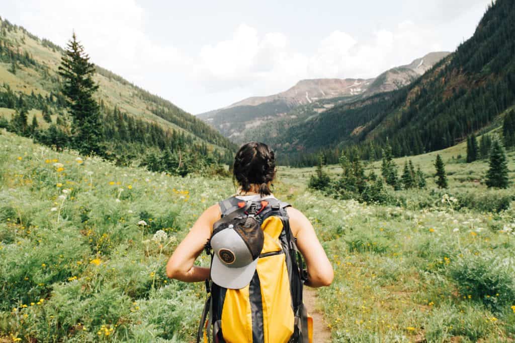What to Wear Hiking: A Complete Guide for Beginners 2 Girl hiking during the summer season wearing a yellow backpack. Wearing short sleeves and her hair braided. Hiking through a green field