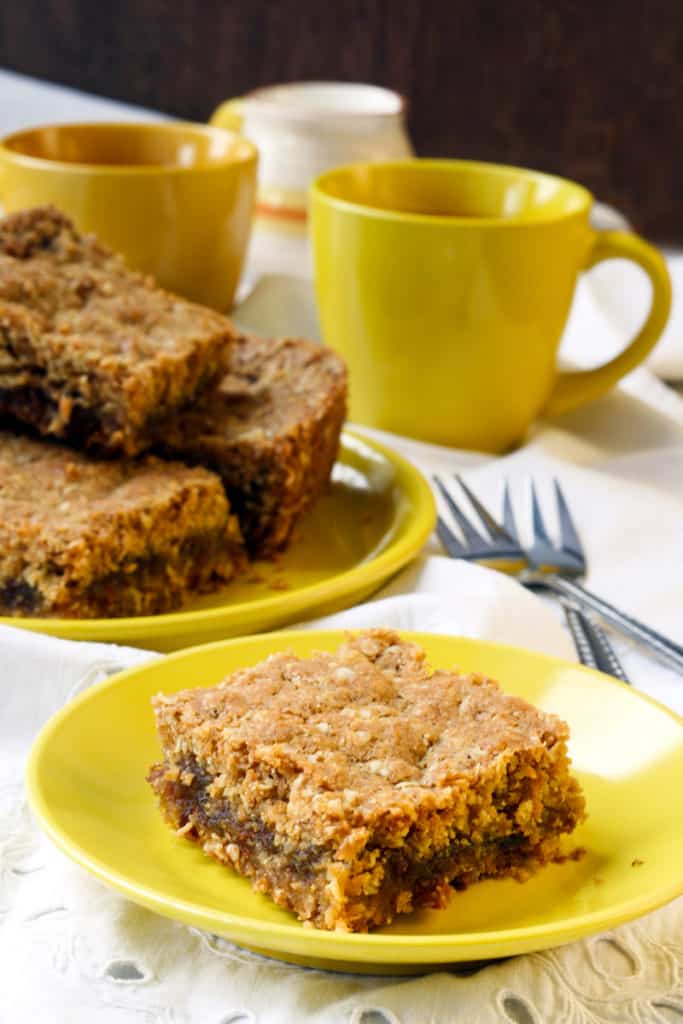 Canada Cuisine to Try: Traditional Food Canada is Famous For 15 Date squares on a yellow plate with yellow mugs in the background