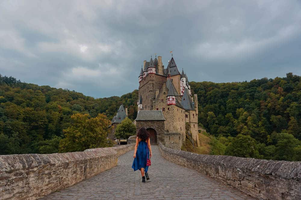 11 Fairytale Castles in Germany: Most Beautiful Castles to Tour 3 Sonal in a blue dress walking along a bridge towards Burg Eltz, a fairytale castle Germany