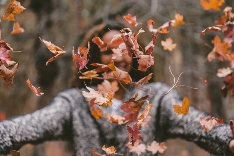 160 Best Fall Quotes and Captions for Instagram 2024 7 Woman throwing autumn leaves in front of the camera and smiling