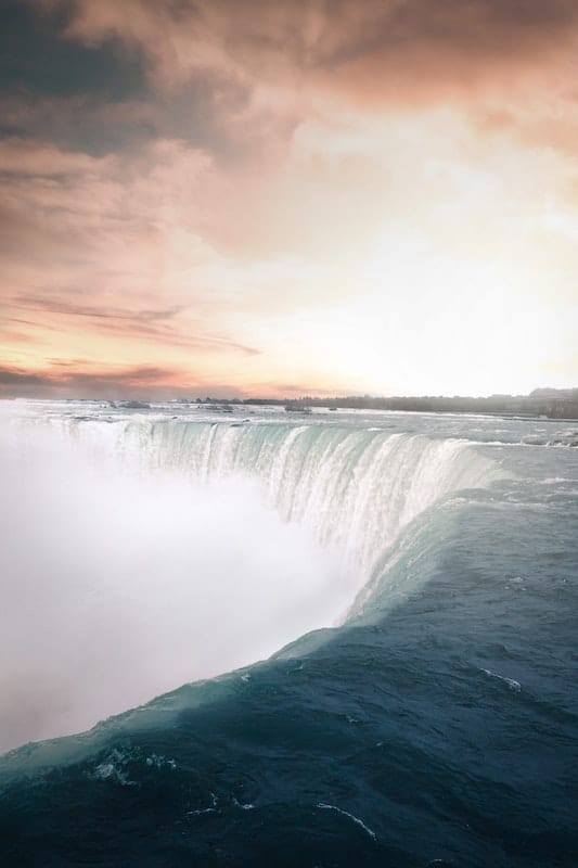 15 Best Niagara Falls Tours from Toronto (2024) 9 A photo of water cascading off a cliff creating a waterfall under an orange-cloudy sky. This view of the Niagara Falls can be seen in the Niagara Falls Tours from Toronto.