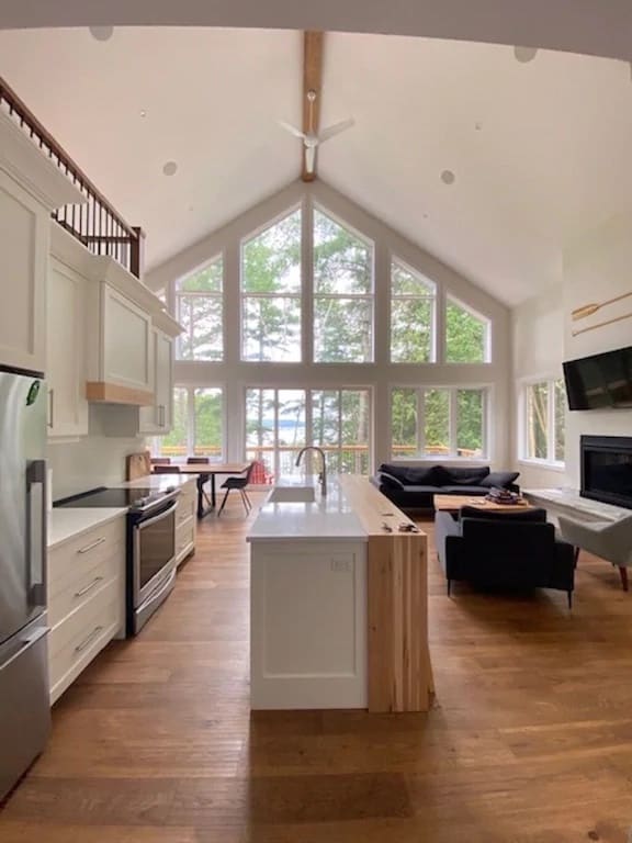 20 Top Airbnb Cottages in Muskoka for an Epic Getaway 28 Heart of 3-Mile Lake cottage. White living room with giant windows and cathedral ceiling. Chef's kitchen and black couches looking at a flat screen TV.