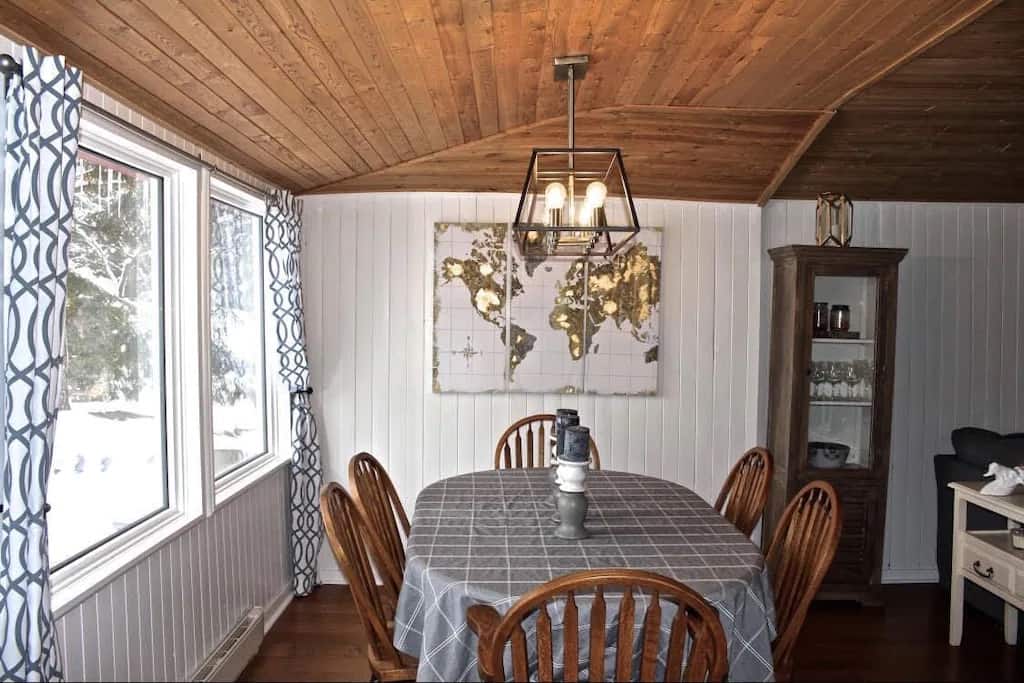 20 Top Airbnb Cottages in Muskoka for an Epic Getaway 9 Dining room of the Charming Lakefront Bala Cottage. Gold decal map on the wall. Wood table with a grey table cloth and big windows on the side.