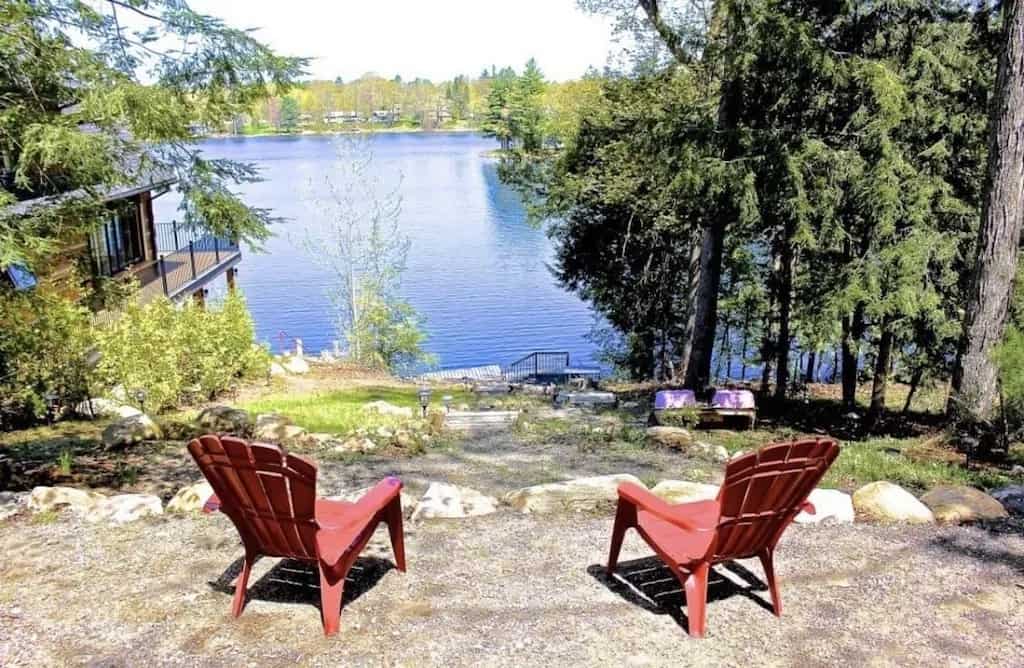 20 Top Airbnb Cottages in Muskoka for an Epic Getaway 7 View of the waterfront from Airbnb cottage Muskoka "Charming Lakefront Bala Cottage". Two red Muskoka chairs overlooking the lake through the trees onto the dock.