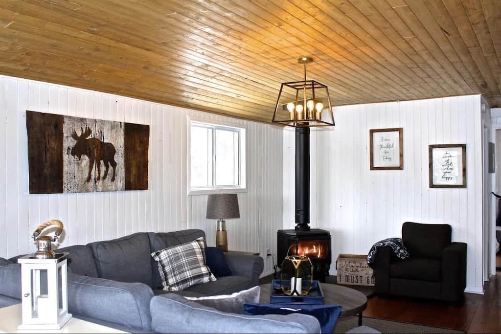 20 Top Airbnb Cottages in Muskoka for an Epic Getaway 8 Living room of the Charming Lakefront Bala Cottage for rent on airbnb. White panelled walls with a wood ceiling, wood burning fireplace, grey couch, and a distressed Canada flag with a moose replacing the maple leaf on the wall.