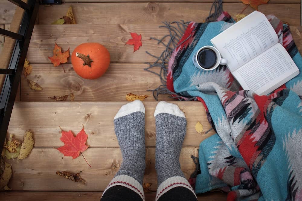 Canada in Autumn: 15 Reasons to Visit + Weather Guide 11 Cozy autumn setting with a book, a cup of coffee, a pumpkin, colorful blanket, and a person wearing socks amidst fallen leaves.