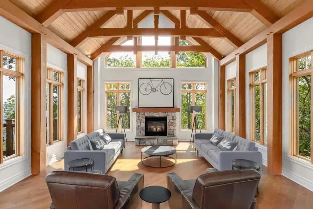 20 Top Airbnb Cottages in Muskoka for an Epic Getaway 49 Infinite Vacations living room. Grey couches and leather arm chairs facing a stone fireplace in a post and beam cottage. White walls with wooden beams and ceiling.