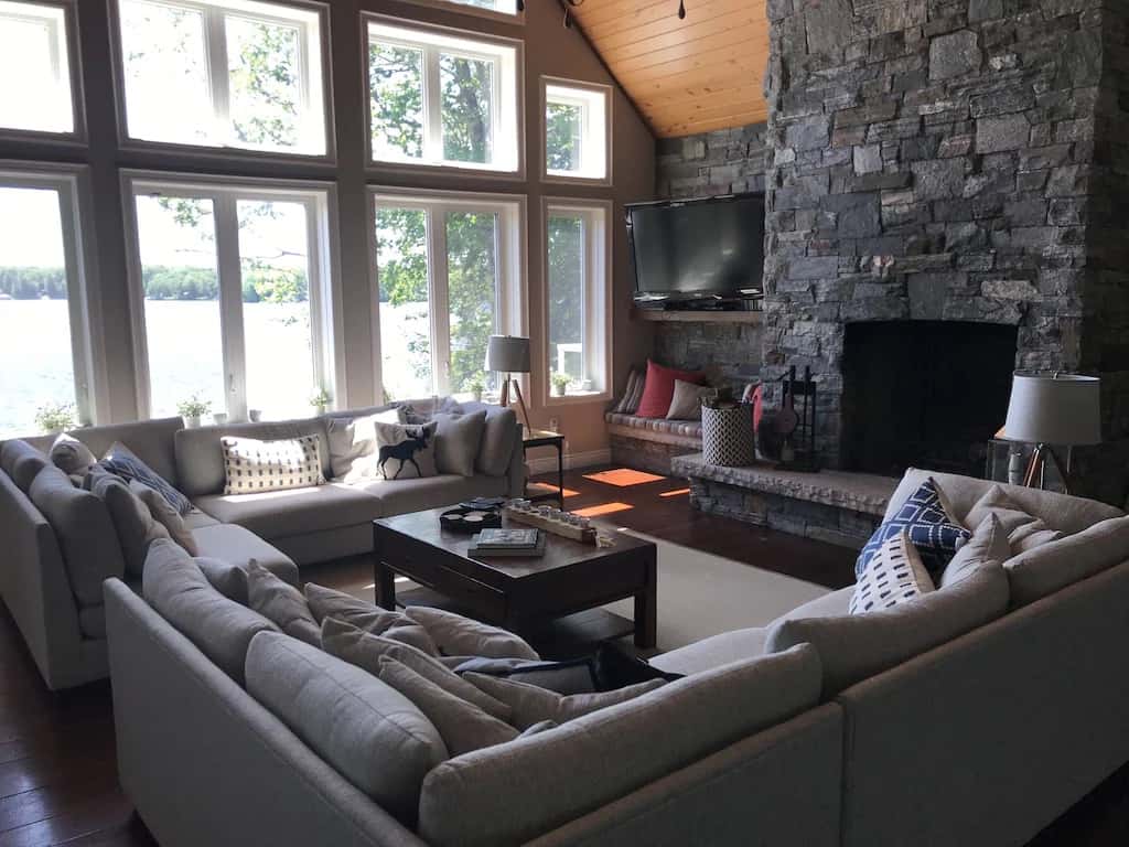 20 Top Airbnb Cottages in Muskoka for an Epic Getaway 62 Stone fireplace in the living room of Muskoka Beach House. Light grey couches and a flat screen TV. Large windows showing the lake and trees.