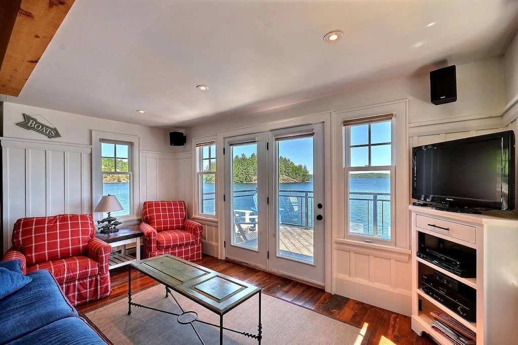 20 Top Airbnb Cottages in Muskoka for an Epic Getaway 33 Muskoka boathouse interior. White walls with red checked arm chairs and a flat screen TV. Doors leading out to patio looking at Lake Muskoka.