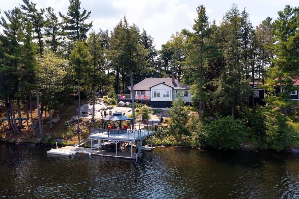 20 Top Airbnb Cottages in Muskoka for an Epic Getaway 41 Exterior view of Gorgeous Lake Muskoka cottage with hot tub. White exterior with brown shaker roof, dock, and elevated patio.