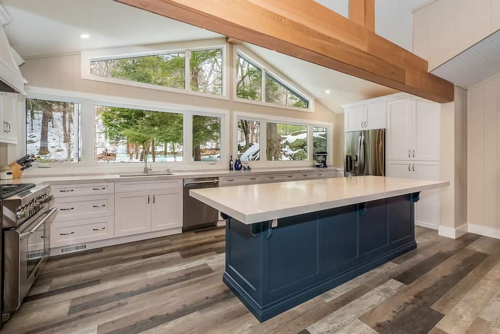 20 Top Airbnb Cottages in Muskoka for an Epic Getaway 43 New Build Lake Rosseau kitchen view of the Airbnb. White marble countertops on a pewter blue island with a pine beam. Large windows looking out into the pine forest.
