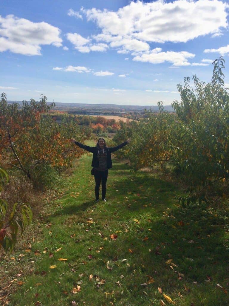 Canada in Autumn: 15 Reasons to Visit + Weather Guide 18 Nina smiling with arms wide in an apple orchard in Annapolis Valley in Canada autumn
