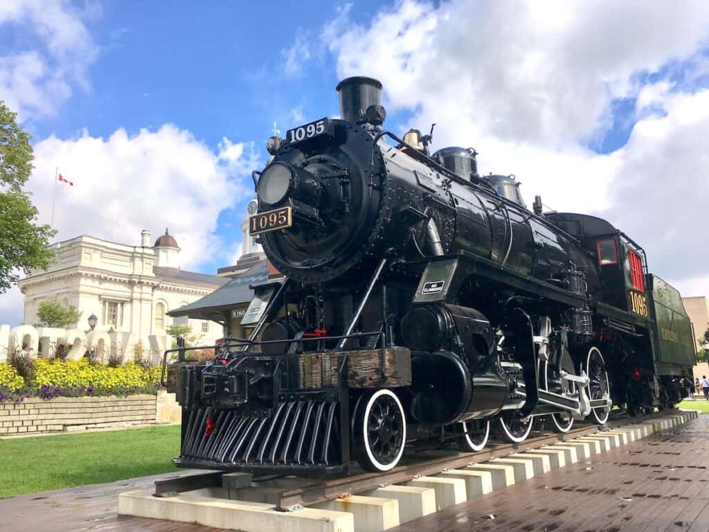 15 Best Cities to Live in Canada in 2024 (Local Tips!) 12 Kingston old train in front of the Kingston town sign and the municipal government building on a sunny day. Kingston is one of the best places to live in Canada for students.