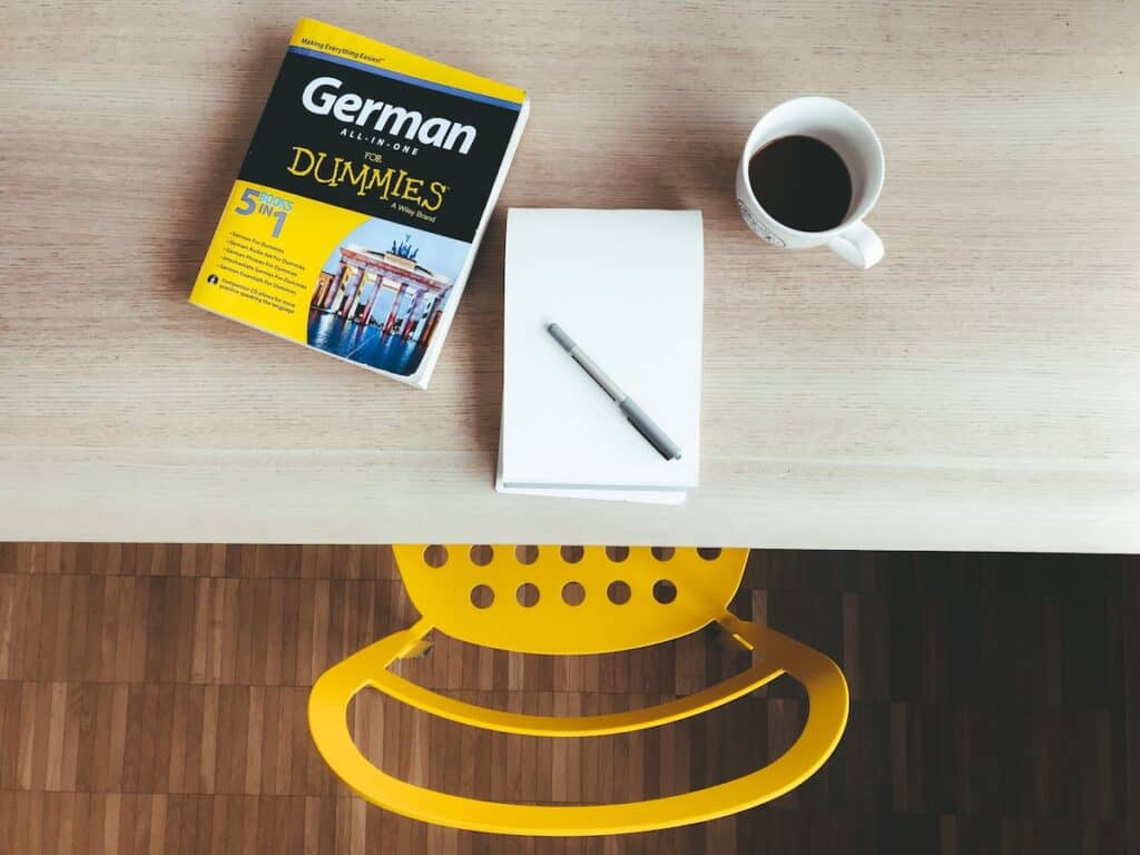 How to Say How Are You in 100 Different Languages 4 Book on learning the German language set on a desk beside a notebook, coffee, and yellow chair.