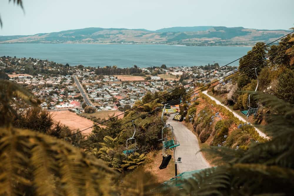 13 Best Places to Live in New Zealand (and Where Not To!) 12 View of the gondola chairs over rotorua that take you up to a viewpoint over the city. You can see the city in the background with small houses.