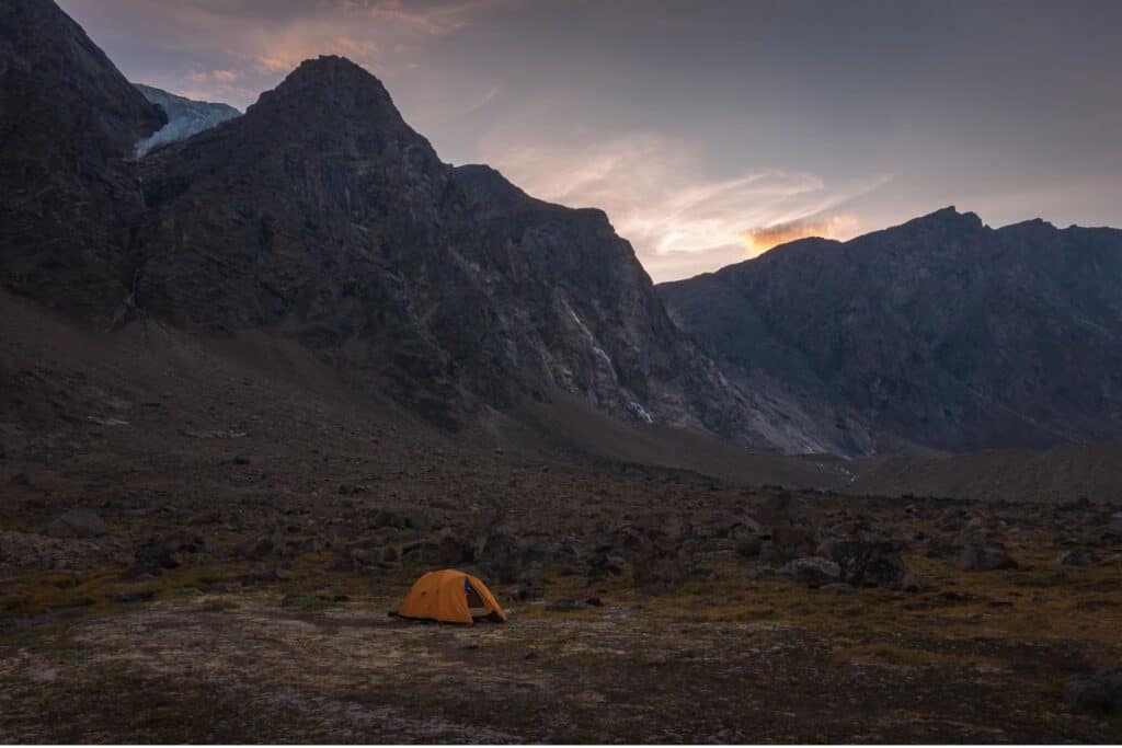 11 Epic Sites for Backcountry Camping in Canada (2024) 6 Yellow tent in Auyuittuq National Park in Canada at twilight with sunset above the mountains.