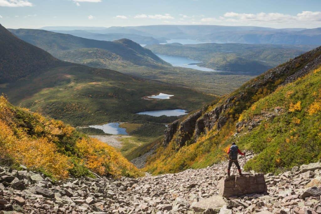11 Epic Sites for Backcountry Camping in Canada (2024) 11 Gross Morne National Park with a person standing in the foregound with a backpack posing. The mountains roll in the distance with small lakes amongst the lichen in Newfoundland.