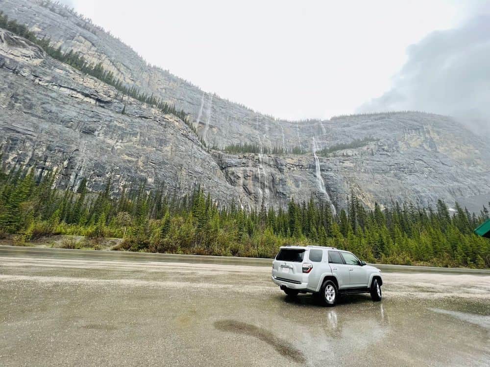 19 Best Stops on the Jasper to Lake Louise Drive 2 Silver SUV parked in front of waterfalls running down the Rocky Mountains