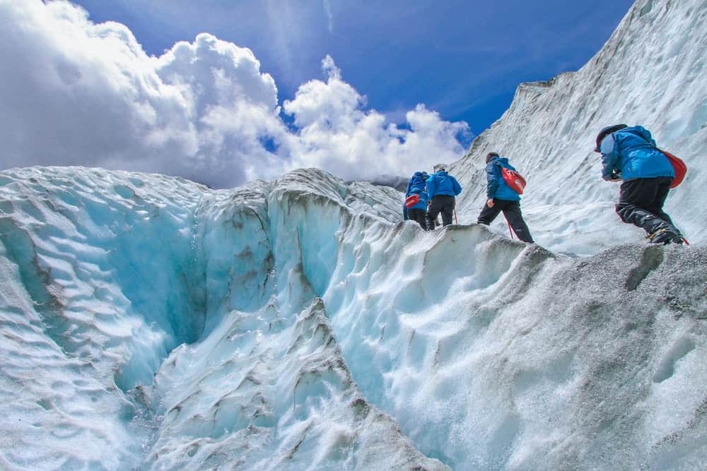 When is winter in New Zealand? (& Should You Visit?) 13 People in blue parkas hiking up the Franz Joseph glacier after landing in a helicopter on a tour.