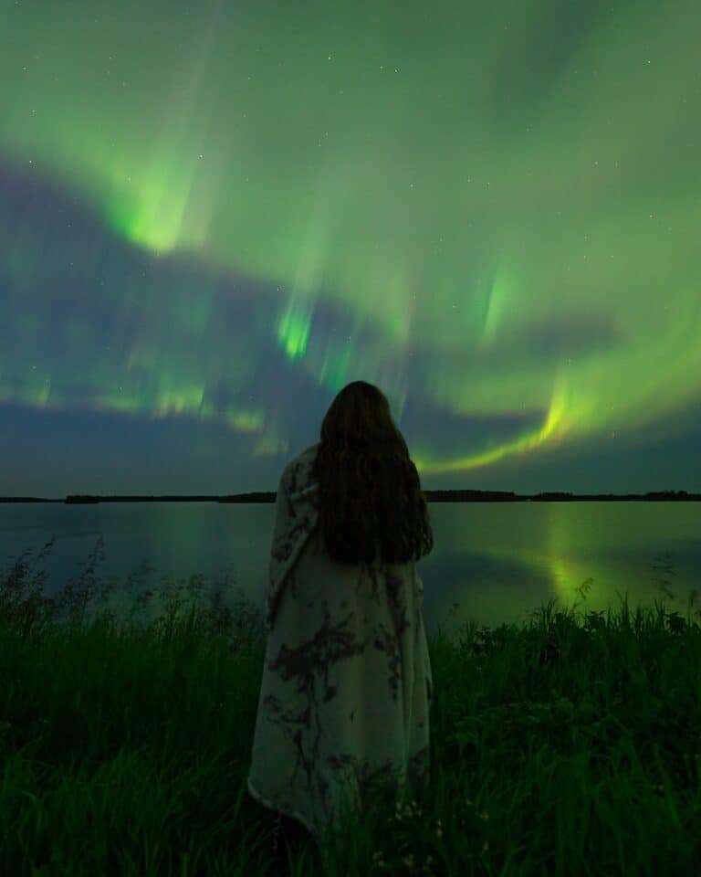 13 Best Canada Northern Lights Tour and When to Go (2024) 33 Woman wrapped in a blanket in front of green northern lights above Saskatchewan in Canada