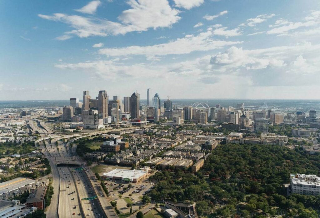 21 Pros and Cons of Living in Dallas in 2024 3 Aerial view of Dallas on a beautiful sunny day featuring its busy roads and tall buildings surrounded by trees.