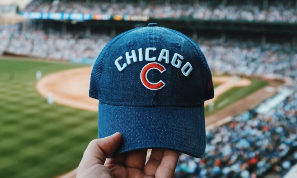 23 Pros and Cons of Living in Chicago in 2024 4 A person holding a blue denim cap of the Chicago baseball team in a baseball stadium surrounded by many people.