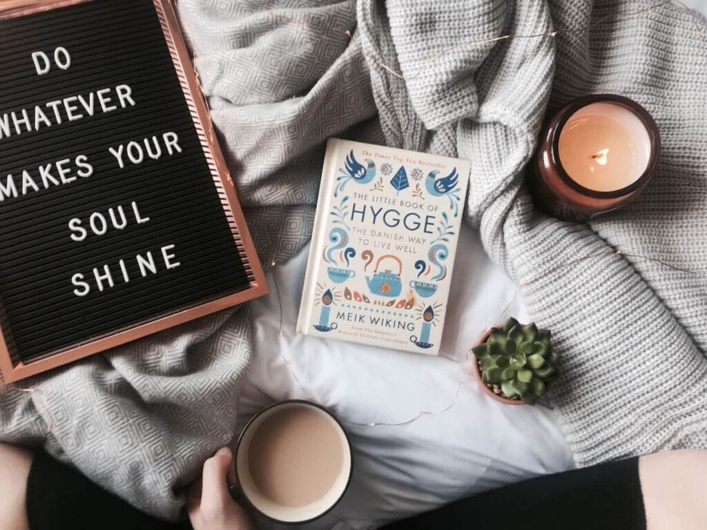 23 Pros and Cons of Living in Denmark in 2024 4 A black letter board with white letters saying "Do whatever makes your soul shine" beside a white book about Hygge, a candle, a succulent and a warm coffee on a white bed in Denmark. Hygge is definitely a pro when living in Denmark.
