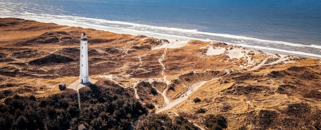 23 Pros and Cons of Living in Denmark in 2024 2 A white lighthouse surrounded by a brown rocky shore in Denmark is near the blue sea.