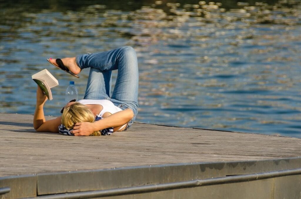 27 Pros and Cons of Living in Vermont in 2024 4 A woman laying down in one of the parks in Vermont reading a book near a lake - you'll have plenty of time to read when you're living in Vermont.