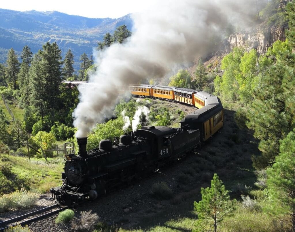 19 Pros and Cons of Living in Colorado in 2024 3 A black steam train on a railroad surrounded by green trees in Colorado.