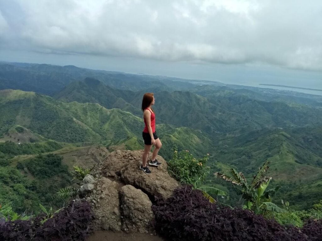 21 Pros and Cons of Living in the Philippines in 2024 3 A woman in a red top standing on top of a rock mountain, surrounded by green mountains in the Philippines - plenty of time to discover the beautiful nature when you're living in the Philippines.