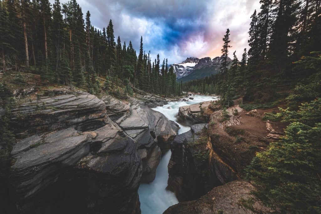 12 Best Jasper Helicopter Tours (2024) 4 A river flowing in between large rocks ina lush green forest. pine trees are surrounding the river underneath a cloudy sky. An exploration hike in this trail is is included in Jasper Helicopter Tours.