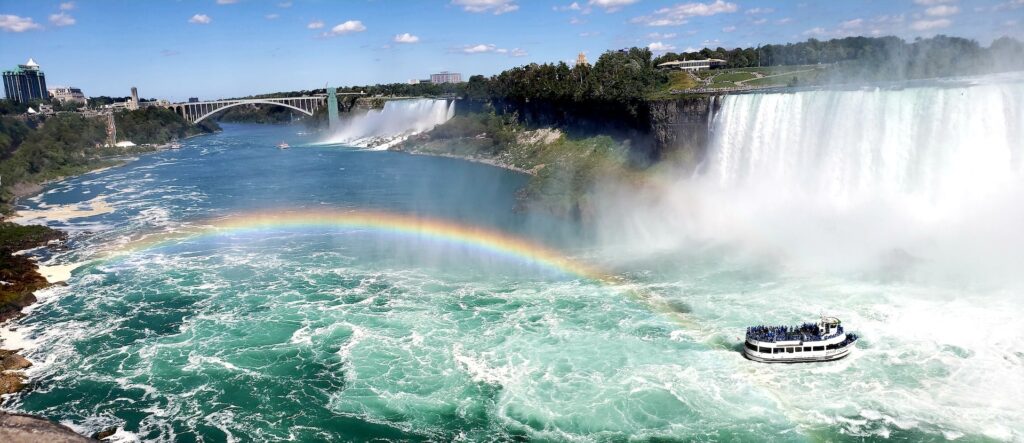 11 Awesome New York Niagara Falls Tours (2024) 9 A black and white ship at the bottom of a waterfall and on one end of a rainbow. A bridge could also be seen near the falls with large trees surrounding it. This is in Niagara Falls where you could have a One Day Tou from New York.