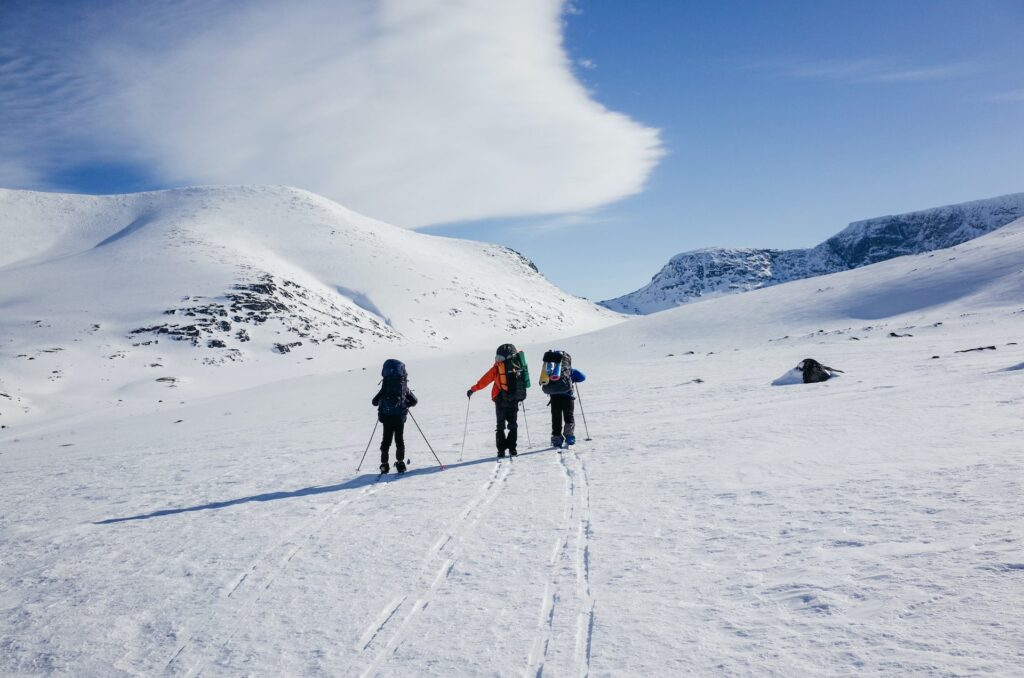 12 Best Jasper Helicopter Tours (2024) 9 Three people with large backpacks skiing in a snow-covered mountain under a blue sky. This is in Jasper Natonal Park's Mountaintop, one of the inclusions for a Jasper Helicopter Tour.