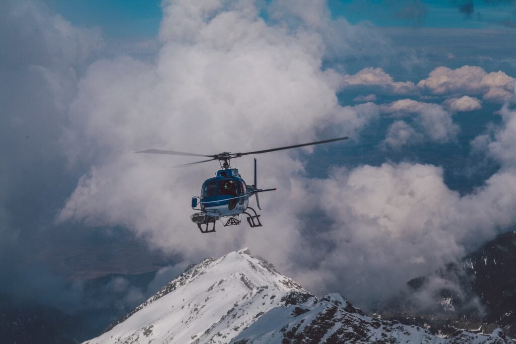 12 Best Jasper Helicopter Tours (2024) 5 A blue helicopter hovering above a snow-covered mountain near the clouds, under a blue sky. This is a helicopter tour in Jasper above the Rocky Mountains.