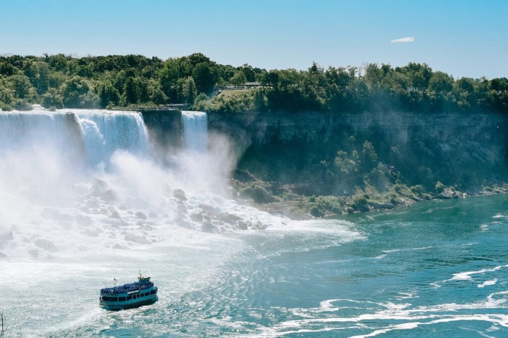 11 Awesome New York Niagara Falls Tours (2024) 7 A white two-deck ship on a clear blue body of water near a waterfall. This boat ride is included in the 1-Day New York Niagara Falls Tour.